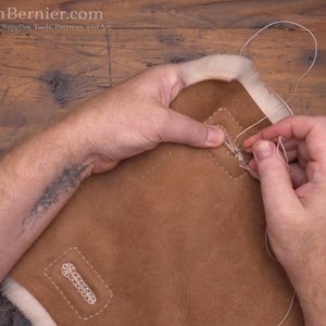May include: Close-up of leatherworking. A person is hand-stitching a brown leather piece with white thread. The leather has a light-colored, fluffy trim. The image includes the website address TonyAllenBernier.com.