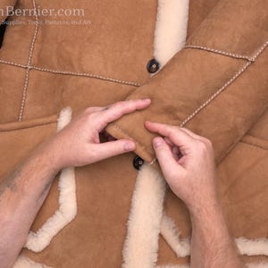 May include: A tan shearling coat with cream-colored trim and stitching. The coat has a button closure and pockets. The sleeves are being held by hands. The coat is displayed on a wooden surface.
