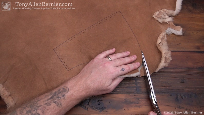 May include: A person cuts a piece of brown leather with a pair of silver scissors. The leather has a pattern drawn on it with a dotted line.