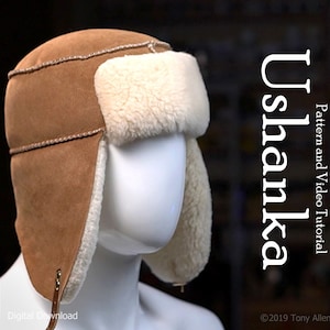 Peut inclure: Un chapeau ushanka en fausse fourrure marron avec des rabats d'oreilles. Le chapeau est doublé de fausse fourrure blanche et possède une sangle en cuir marron avec une boucle en métal. Le texte "Ushanka" est écrit verticalement en blanc sur le côté droit de l'image. Le texte "Pattern and Video Tutorial" est écrit en blanc sur le côté droit de l'image. Le texte "©2019 Tony Allen Bernier" est écrit en blanc en bas à droite de l'image.