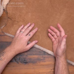 May include: A close-up of a tanned leather hide with a white, fluffy edge. The hide is being held by two hands. The leather has a smooth texture and is a warm brown color. The image is taken from a top-down perspective.