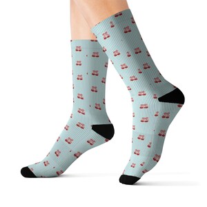 May include: A pair of light blue crew socks featuring a repeating pattern of small red cowboy boots. The socks have a ribbed texture throughout the fabric, with solid black fabric at the toe and heel sections for a distinct contrast in the design.