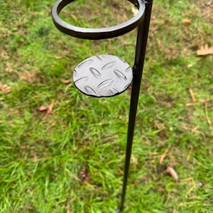 May include: A black metal plant stand with a circular top and a small, round, textured platform. The stand is designed to be placed in the ground, with a long, slender pole. The stand is set against a backdrop of green grass.