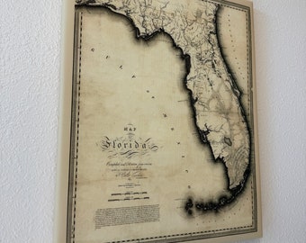 May include: An antique map of Florida, printed on aged paper with a cream background. The map shows the state's coastline, rivers, and major cities. The title "MAP of FLORIDA" is printed in a decorative script at the top of the map. The map is surrounded by a border with a decorative pattern.