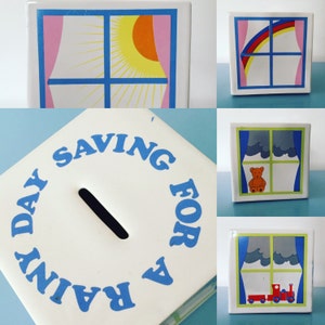 Puede incluir: Una alcancía de cerámica blanca con un círculo azul alrededor de las palabras "Saving for a Rainy Day". La alcancía tiene una ranura para monedas y una ventana con una imagen de un cielo nublado.