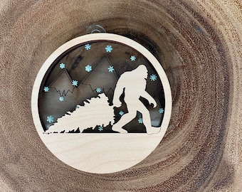 Könnte beinhalten: Ein hölzerner Weihnachtsbaumschmuck mit der Silhouette eines Bigfoots, der einen Weihnachtsbaum trägt. Der Schmuck ist mit blauen Glitzer-Schneeflocken auf weißem Hintergrund verziert.