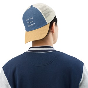 Puede incluir: Una gorra de béisbol azul y tostada con una malla blanca en la parte posterior. La parte delantera de la gorra tiene el texto "Your dog speaks Chinese?!" bordado en blanco.