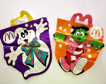 May include: Two Halloween-themed reusable shopping bags. One bag is purple with a white ghost wearing a pink, yellow, and blue bow. The other bag is orange with a green monster wearing a pink and white striped shirt. Both bags have a yellow "M" logo and the text "Boo Boo" on them.