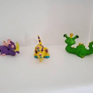 May include: Three small plastic figurines of fantastical creatures. A purple dragon with yellow horns, a yellow creature with multiple eyes and a green creature with a long tail.