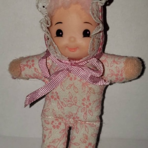 May include: A small pink and white fabric doll with a pink bow and a white lace bonnet. The doll has a painted face with black eyes and a pink smile.