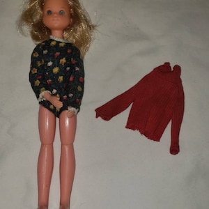 May include: A vintage doll with blonde hair, blue eyes, and a floral print dress. The doll is wearing a red sweater.