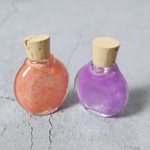 May include: Two small, clear glass bottles with cork stoppers. One bottle contains an orange-pink substance, the other a purple substance. The bottles have a rounded shape and sit on a light gray surface.
