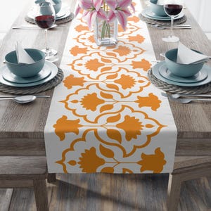 May include: A white table runner featuring a repeating orange floral design. The runner is laid on a wooden table set for a meal, with plates, bowls, cutlery, and wine glasses. A vase of pink flowers is in the centre.