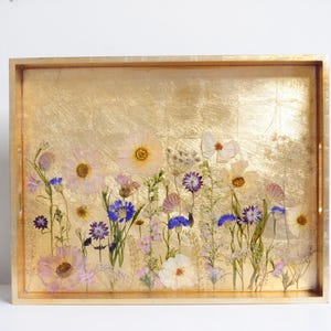 May include: A gold tray with a pressed flower design. The tray features a variety of flowers in shades of pink, white, and blue, including cosmos, daisies, and wildflowers. The flowers are arranged in a random pattern on a gold background.