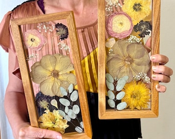 May include: Two rectangular wooden frames showcasing pressed flowers. The frames contain a variety of dried flowers in shades of yellow, pink, and white, with green foliage. The frames are held up by a person.