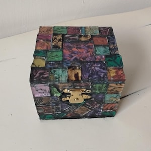 May include: A small, decorative box with a mosaic-like design. The box is covered in small, irregularly shaped tiles in shades of purple, green, brown, and gold. It has a gold-colored clasp and hinges. The box is likely used for storing small items.