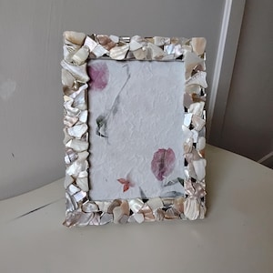 May include: Rectangular photo frame with a mosaic border made of iridescent shell pieces. The frame surrounds a piece of paper with a floral design in shades of pink and gray. The frame is on a white surface.