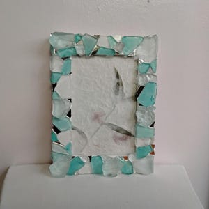 May include: A rectangular picture frame decorated with a mosaic of translucent sea glass pieces in shades of aqua, turquoise, and clear white. The frame surrounds a textured, off-white paper insert featuring a faint, delicate botanical print.