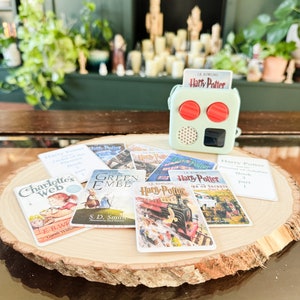 May include: A collection of miniature book covers featuring popular children's and young adult fiction titles, including "Charlotte's Web", "Green Ember", and "Harry Potter". The covers are arranged on a wooden slice, with a small green and red radio-like device in the background.