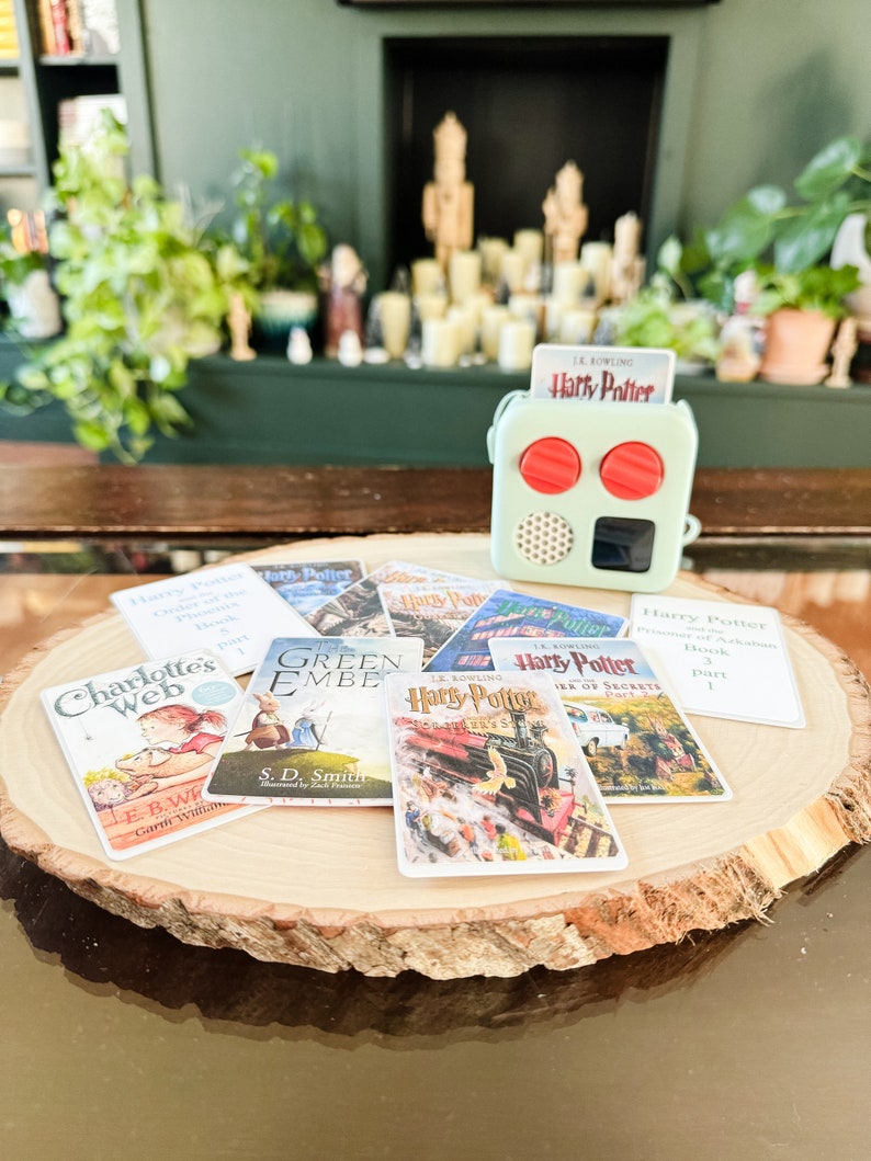 May include: A collection of miniature book covers featuring popular children's and young adult fiction titles, including "Charlotte's Web", "Green Ember", and "Harry Potter". The covers are arranged on a wooden slice, with a small green and red radio-like device in the background.