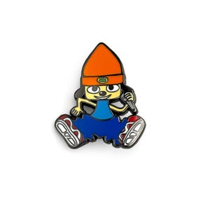 Puede incluir: Un colorido pin de esmalte con un personaje de dibujos animados con cara amarilla, gorro naranja, atuendo azul y zapatillas rojas. El personaje sostiene un micrófono y tiene un contorno negro. El pin está sobre un fondo blanco.