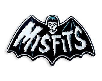 Puede incluir: Pin de esmalte negro y blanco con el logotipo de la banda Misfits. El logotipo es un murciélago con el nombre de la banda escrito en letras blancas a través de las alas. Una calavera con una cara azul está en el centro del murciélago.