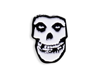 Könnte beinhalten: Eine schwarz-weiße Emaille-Anstecknadel mit einem Schädel mit einem breiten Lächeln. Der Schädel hat ein weißes Gesicht und schwarze Augen, Nase und Mund.