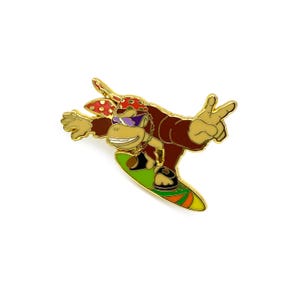 May include: A gold-colored pin featuring a cartoon monkey wearing a red bandana with white polka dots and sunglasses, riding a green and yellow surfboard.