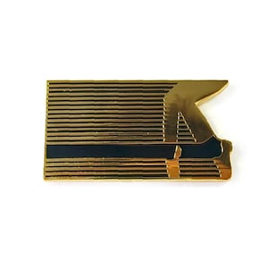 May include: A gold-colored pin with a black stripe and a stylized letter "A" design. The pin features horizontal lines and a geometric shape, creating a modern and abstract aesthetic. The pin is likely made of metal and has a shiny finish.
