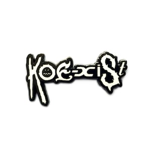 May include: Black and white enamel pin with the word "KOECIST" in a stylized font. The "O" in "KOECIST" is replaced with a smiley face.