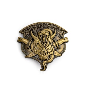 May include: A bronze-colored metal emblem with the word "DREADNOKS" arched across the top. The emblem features a skull with horns, a snake, and crossed swords. The design has a detailed, embossed appearance.