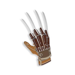 May include: A silver and brown metal pin depicting a glove with four long blades attached to the fingers. The glove is brown with a tan wrist strap.
