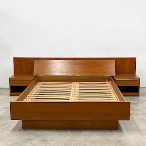 May include: A teak wood bed frame featuring an integrated headboard with two attached nightstands, each containing a single drawer and an open storage shelf. The bed frame includes a slatted base and a wide, flat footboard in a warm brown finish.