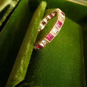 May include: A gold band ring with a row of square-shaped ruby and diamond stones.