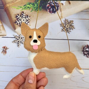 May include: A felt corgi dog decoration with a tan body, white chest and paws, and a pink tongue. The dog has black eyes and a white stripe on its face. A gold string is attached for hanging. Christmas decoration.
