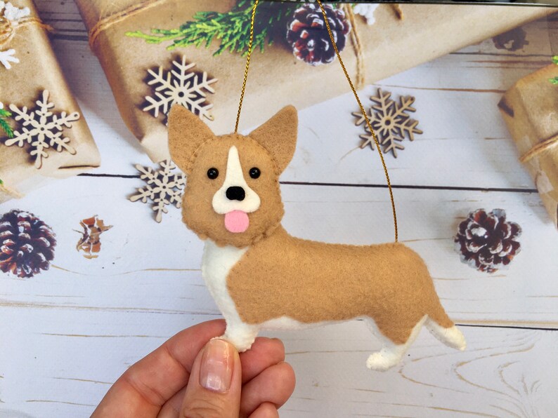 May include: A felt corgi dog decoration with a tan body, white chest and paws, and a pink tongue. The dog has black eyes and a white stripe on its face. A gold string is attached for hanging. Christmas decoration.
