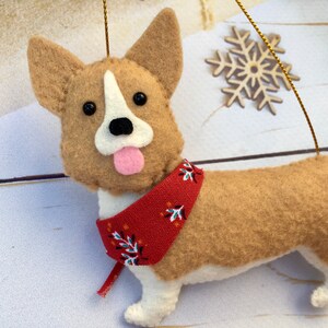 May include: A felt corgi dog decoration with a red bandana featuring a white and blue floral pattern. The dog has tan fur, white accents, and a pink tongue. A gold string is attached for hanging.