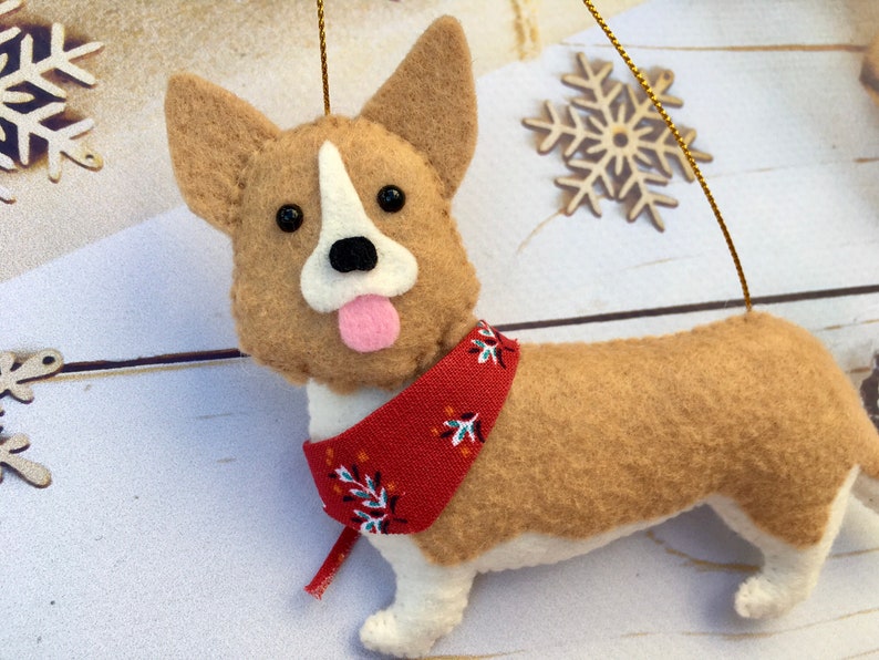 May include: A felt corgi dog decoration with a red bandana featuring a white and blue floral pattern. The dog has tan fur, white accents, and a pink tongue. A gold string is attached for hanging.