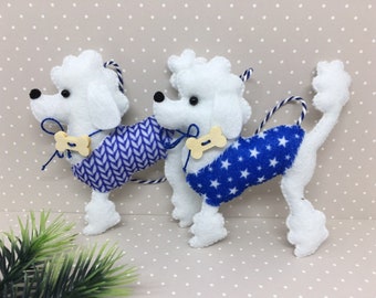 Puede incluir: Dos adornos de caniche de fieltro blanco con chaquetas estampadas en azul y blanco y detalles en forma de hueso. Una chaqueta tiene un patrón de chevrón, la otra estrellas blancas. Cada uno tiene una correa de cordón azul y blanco.
