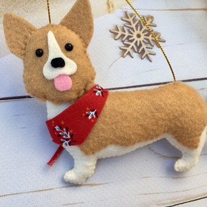May include: A felt corgi dog decoration with a red bandana. The dog has tan and white fur, black eyes, and a pink tongue. A gold string is attached for hanging. Decorative snowflakes are in the background.