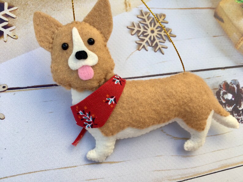 May include: A felt corgi dog decoration with a red bandana. The dog has tan and white fur, black eyes, and a pink tongue. A gold string is attached for hanging. Decorative snowflakes are in the background.