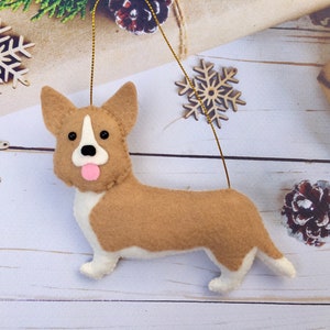 May include: A felt corgi dog decoration with a gold hanging string. The ornament is tan and white with black eyes and a pink tongue. Wooden snowflakes and a pine cone are in the background.