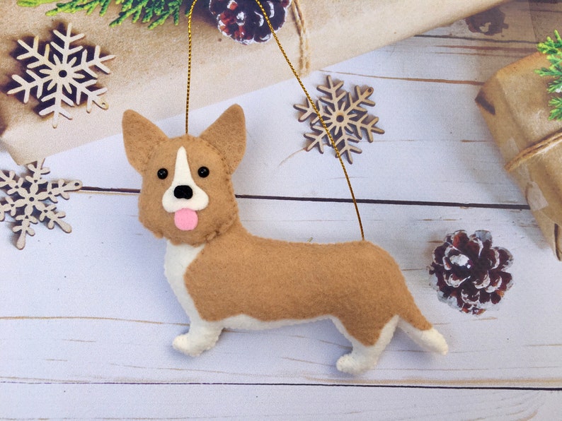 May include: A felt corgi dog decoration with a gold hanging string. The ornament is tan and white with black eyes and a pink tongue. Wooden snowflakes and a pine cone are in the background.