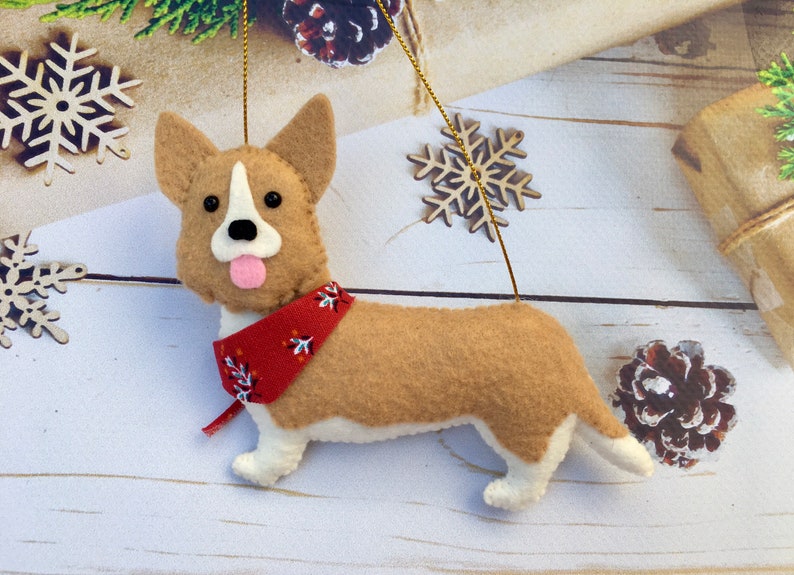 May include: A felt corgi dog decoration with a red bandana. The dog has brown and white fur, black eyes, and a pink tongue. The ornament is suspended by a gold string and is a festive holiday decoration.