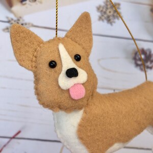 May include: A felt corgi dog decoration with tan and white fur, black eyes, and a pink tongue. It has a gold hanging string. This ornament is suitable for Christmas or other festive occasions.
