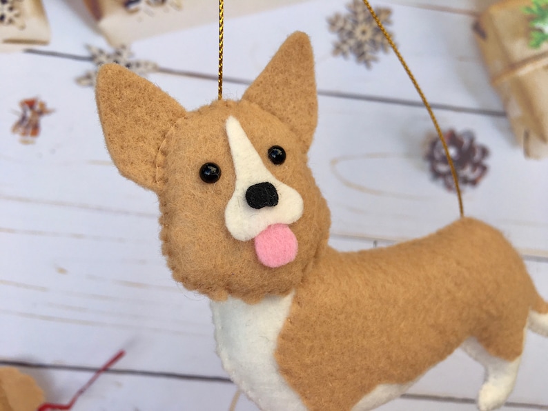 May include: A felt corgi dog decoration with tan and white fur, black eyes, and a pink tongue. It has a gold hanging string. This ornament is suitable for Christmas or other festive occasions.
