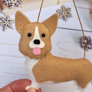 May include: A felt corgi dog ornament. The dog has a tan body, white chest and legs, and a pink tongue. It has black eyes and a white stripe on its face. A gold string is attached for hanging.