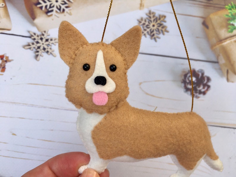 May include: A felt corgi dog ornament. The dog has a tan body, white chest and legs, and a pink tongue. It has black eyes and a white stripe on its face. A gold string is attached for hanging.