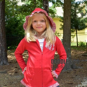 May include: A young girl wearing a red hooded zip-up jacket with a white and red gingham trim and hood lining. The jacket has a white monogram on the chest and is paired with dark blue leggings. The text "DOODLE & CO DESIGNS" is visible.