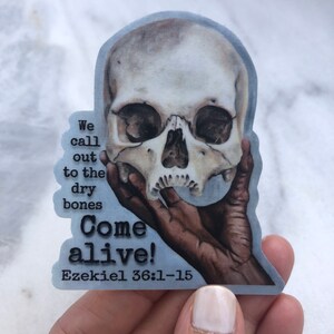 May include: A sticker with a hand holding a human skull. The sticker has the text "We call out to the dry bones Come alive! Ezekiel 36:1-15" on it.
